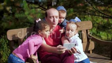 一个带孩子的男人坐在长凳上的智能手机上拍照。 幸福的一家人在公园里自拍。 家庭